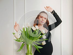 Pregnant woman with a hairstyle and makeup on her face in a black tight dress with white tulips