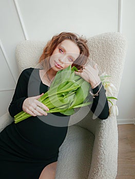 Pregnant woman with a hairstyle and makeup on her face in a black tight dress with white tulips