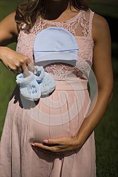 Pregnant woman hand holding blue booties and cap for the newborn baby touching her stomach