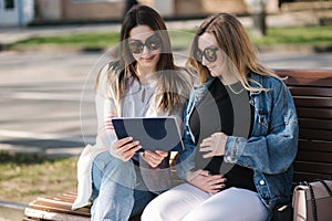 Pregnant woman with her best friend sitting in the park and looking for baby stroller using tablet. Two females in