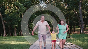 Pregnant woman with her family walking in the park along the path holding the little girl to the hands and raising her