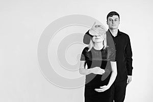 Pregnant woman and her husband covers her face with her hand in black clothes on a white background. Black and white picture.