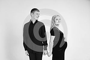 Pregnant woman and her husband holding hands in black clothes on a white background. Black and white picture. Beautiful married