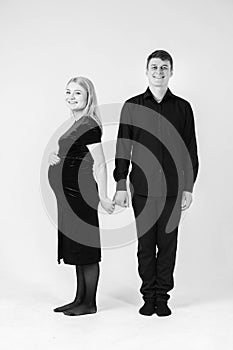 Pregnant woman and her husband holding hands in black clothes on a white background. Black and white picture. Beautiful married