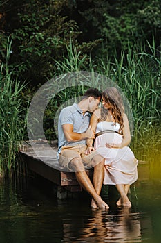 Pregnant woman and her husband sitting at a lake while he is kissing her shoulder gently