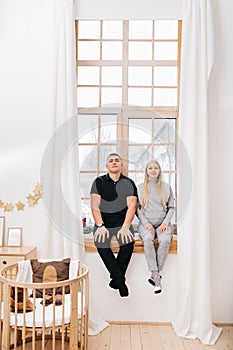 A pregnant woman and her husband are waiting for the baby and sitting on a large panoramic window. wooden cot is in the nursery.