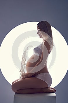 Pregnant woman in her last month of pregnancy in her underwear sits on a cube against a white glowing circle. Waiting for the