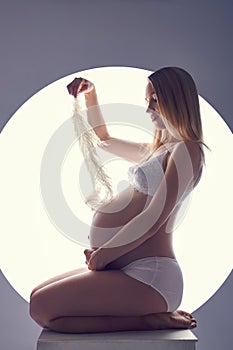 Pregnant woman in her last month of pregnancy in her underwear sits on a cube against a white glowing circle. Waiting for the