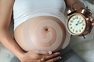 Pregnant woman holding an alarm clock on tummy sitting on sofa. Waiting baby concept