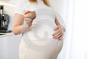 A pregnant woman is holding an apple and put her hand on her stomach.