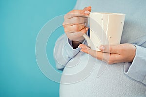 Pregnant woman holding ceramic cup of tea or coffee on belly. Young girl in blue wearing expecting baby. Maternity