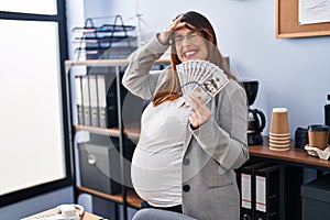 Pregnant woman holding dollars at the office stressed and frustrated with hand on head, surprised and angry face