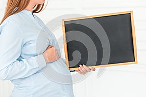 Pregnant woman is holding an empty blackboard. Close-up, copy space, indoors