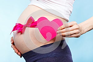 Pregnant woman holding heart and touching her belly with pink ribbon, concept of expecting for newborn girl