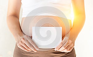 Pregnant woman holding her hands on the belly and holding a blank white pape