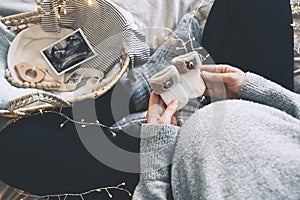 Pregnant woman holding little baby socks near her belly. Expectant mother preparing wicker basket with ultrasound image and stuff