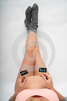 Pregnant woman holding mini blackboards with written boy and girl while laying on her bed with legs on the wall at home