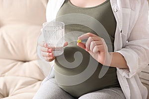 Pregnant woman holding pill and glass with water on sofa, closeup