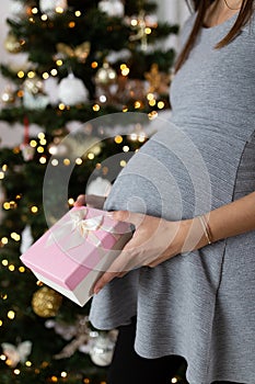 Pregnant woman holding rose christmas present next to her belly with christmas tree in the background