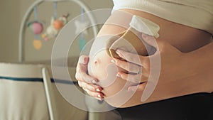 Pregnant woman holding tiny baby socks over her belly. Baby crib with spinning toys in the backgournd. Expecting and