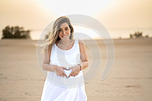 Pregnant woman holding tummy and showing heart shape with hands at beach.