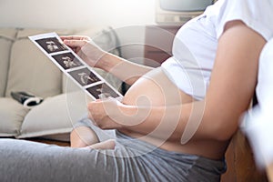 Pregnant woman holding ultrasound scan photo on her belly at home