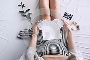 Pregnant woman holding white baby bodysuit preparing to child birth during pregnancy