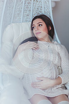 Pregnant woman holds her belly. Wears white knitted sweater. White effect photo