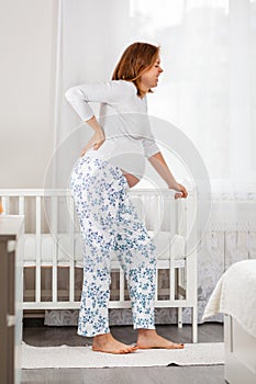 A pregnant woman holds her hands on the small of her back and leans on a baby cradle. Children`s room in the background. The