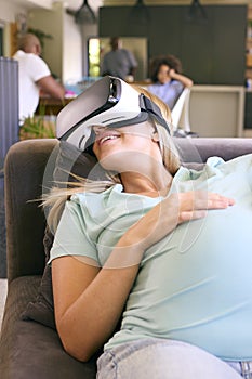 Pregnant Woman At Home Lying On Sofa Wearing VR Headset
