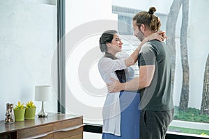 Pregnant woman hug and entwine her husband and stand in living room with glass window that show some part of her home garden. They