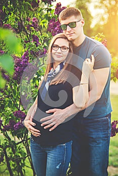 Pregnant woman and husband in nature park, blooming lilac