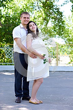 Pregnant woman with husband posing in the city park, family portrait, summer season, green grass and trees