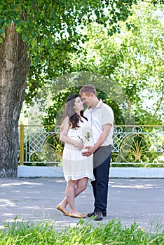 Pregnant woman with husband posing in the city park, family portrait, summer season, green grass and trees