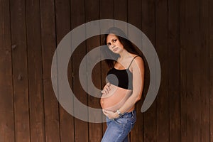 Pregnant woman in jeans and black top holds hands on belly on a dark brown background. Pregnancy, maternity, preparation and