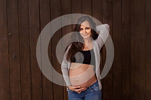 Pregnant woman in jeans and black top holds hands on belly on a dark brown background. Pregnancy, maternity, preparation and