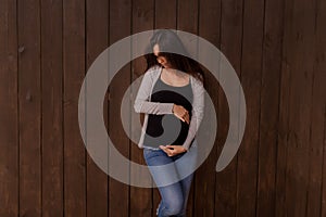 Pregnant woman in jeans and black top holds hands on belly on a dark brown background. Pregnancy, maternity, preparation and