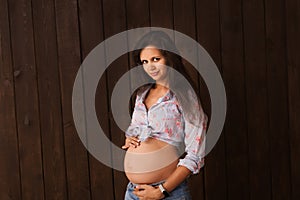 Pregnant woman in jeans shorts and blue top holds hands on belly on a dark brown background. Pregnancy, maternity, preparation and