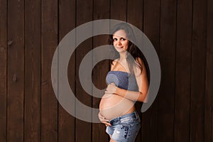 Pregnant woman in jeans shorts and blue top holds hands on belly on a dark brown background. Pregnancy, maternity, preparation and