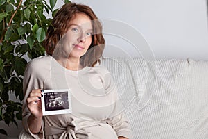 Pregnant woman keeping hand on belly and holding ultrasound image at home interiors. Pregnancy, parenthood, preparation