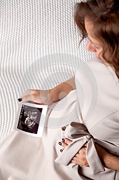 Pregnant woman keeping hand on belly and holding ultrasound image at home interiors. Pregnancy, parenthood, preparation