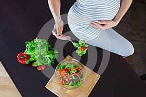 Pregnant woman in kitchen is eating vegetable salad.