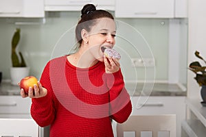 Pregnant woman in kitchen holding apple in hand and eating cake. Young attractive female decides to eat unhealthy food, looks