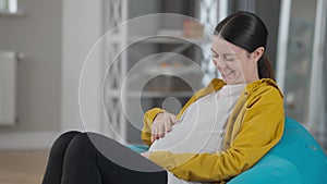 Pregnant woman laughing as baby kicking in belly. Positive smiling Caucasian expectant enjoying leisure at home indoors