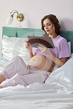 A pregnant woman lies in bed of her domestic bedroom. Female reads an e-book