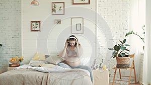 Pregnant woman listening music in bed. Expectant mother sitting with headphones.