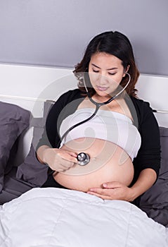 Pregnant woman listening to her belly with a stethoscope on bed