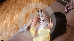 A pregnant woman is looking at ultrasound scans while sitting on the couch. Anticipating the birth of a child. Family