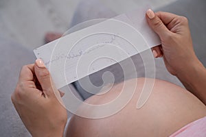 A pregnant woman looks at the results of cardiotocography while sitting on the couch. Close up of the belly.