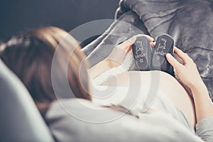 Pregnant woman lying on the sofa and holding shoes unborn child.